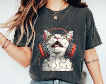 May include: Dark gray t-shirt featuring a graphic of a happy cat wearing red and blue headphones. The cat has its mouth open and eyes closed. The shirt is a casual style.
