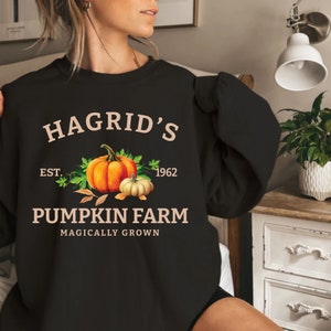 May include: Black sweatshirt with the text "Hagrid's Pumpkin Farm" and an illustration of two pumpkins. The design includes the year "1962" and the phrase "Magically Grown". The pumpkins are orange and white.