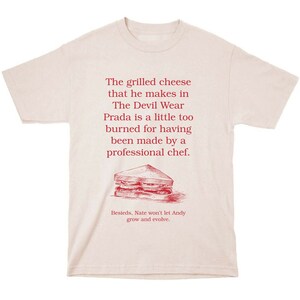 May include: A light pink t-shirt with a red graphic print of a grilled cheese sandwich and text that reads: "The grilled cheese that he makes in The Devil Wear Prada is a little too burned for having been made by a professional chef. Besieds, Nate won't let Andy grow and evolve."