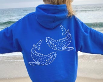 May include: Royal blue hoodie with a white whale design on the back. The design features two whales with dotted patterns. The hoodie has a hood and long sleeves. The background shows a beach and ocean.
