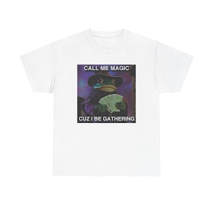 May include: White t-shirt featuring a graphic of a frog dressed as a wizard, holding money. The image includes the text "CALL ME MAGIC CUZ I BE GATHERING". The frog wears a black hat and robe against a purple and green background.