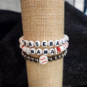 May include: Three beaded bracelets with the words "BASEBALL MAMA". The top bracelet has white beads with black letters and red accents. The middle bracelet has white beads with black letters and silver accents. The bottom bracelet has dark gray beads with a baseball charm.
