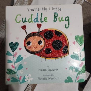 Puede incluir: Un libro infantil titulado "You're My Little Cuddle Bug". La portada presenta una ilustración de mariquita brillante con acentos rojos, negros y beige. El título está en purpurina verde y azul. Autora: Nicola Edwards, ilustradora: Natalie Marshall.