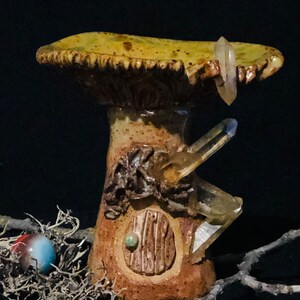 Op de afbeelding: Een keramische paddenstoelvormige wierookhouder met een kleine deur en een kristal erop. De houder is versierd met bruine en groene glazuur.