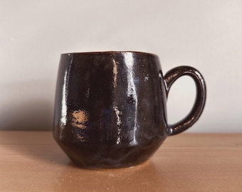 May include: A dark brown ceramic mug with a rounded body and a handle. The mug has a glossy finish and a slightly textured surface.
