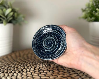 May include: A small, round, dark blue ceramic dish featuring a textured, spiral pattern of raised lines radiating from the center. The surface has a glossy finish with white highlights accentuating the intricate, carved-like grooves on the plate.