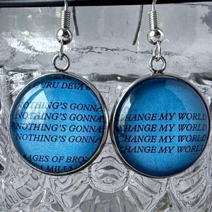 May include: A pair of silver earrings with blue glass round charms. The charms have lyrics from the song "Nothing's Gonna Change My World" by  Guru Deva printed on them.