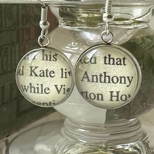May include: Silver earrings with glass charms featuring text from a book. The left earring says "This is Kate living while Victoria", and the right earring says "I said that Anthony Horton How".