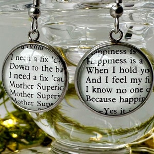 May include: A pair of silver dangle earrings with round glass charms. The charms feature black text on a white background. The left charm reads "I need a fix Down to the ba I need a fix 'cau Mother Superic Mother Super Mother Sup". The right charm reads "Happiness is happiness is a When I hold yo And I feel my fi I know no one q Because happin Yes it".