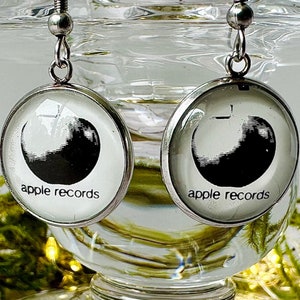 May include: A pair of silver earrings with round glass charms featuring the Apple Records logo in black and white. The charms have the text "apple records" printed on them.