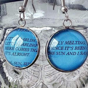 May include: A pair of silver dangle earrings with blue glass cabochons. The glass cabochons have text printed on them. The text on the left earring reads "Little darling, little darling, here comes the, it's alright." The text on the right earring reads "Only melting, since it's been, the sun and I say."