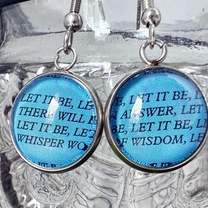 May include: A pair of silver earrings with blue glass charms. The charms have the lyrics "Let it be, let there will be, let it be, let it be, whisper words of wisdom, let it be" printed on them.