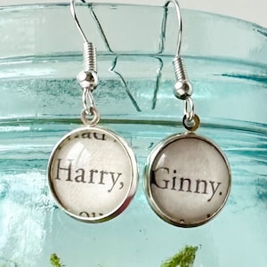 May include: Silver-toned dangle earrings with circular charms. Each charm features text from a book page, one reading "Harry," the other "Ginny." The earrings hang in front of a blue glass jar filled with green moss.