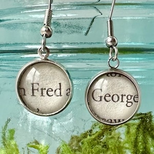 Puede incluir: Un par de pendientes de plata con colgantes de vidrio. Los colgantes son redondos y presentan los nombres "Fred" y "George" impresos en texto negro sobre fondo blanco.