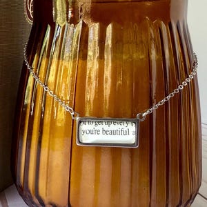 May include: A silver-toned necklace with a rectangular pendant. The pendant features text that reads "to get up every day you're beautiful". The necklace is displayed against a brown, ribbed glass jar.