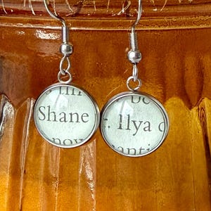 May include: A pair of round earrings with a silver-colored metal frame. Each earring features a circular design with text from a book page. One earring reads "Shane" and the other reads "Ilya". The earrings hang from silver-colored hooks.