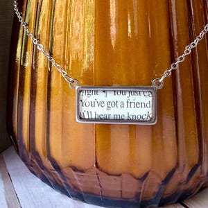 May include: A silver-toned necklace with a rectangular pendant. The pendant features text from a book page: "You've got a friend, I'll hear me knock." The necklace is displayed against a blurred, amber-colored background.