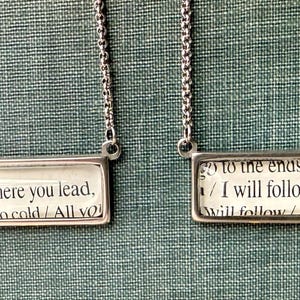 May include: Two silver-toned necklaces with rectangular pendants. Each pendant has a printed text design. One reads "Where you lead," and the other reads "to the ends of the earth / I will follow."