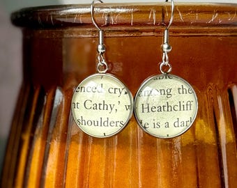 May include: Silver-toned dangle earrings with round, clear glass pendants. Each pendant features text from a book page, with visible words like "Cathy" and "Heathcliff". The earrings hang from a silver hook.