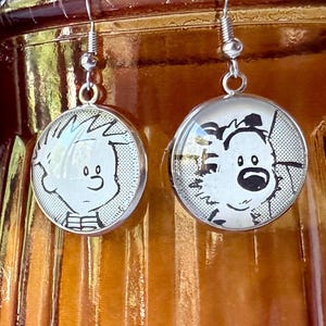 May include: A pair of silver-toned dangle earrings. Each earring features a round, clear glass cabochon with a black and white cartoon image of Calvin and Hobbes characters. The earrings have a simple hook closure.