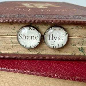 May include: Two round, silver-toned stud earrings with the names "Shane" and "Ilya" printed on a white background. The earrings are set against a vintage book with a red cover and aged pages.