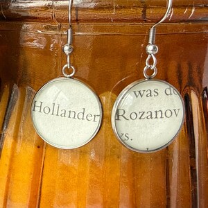 May include: Silver-toned dangle earrings with circular pendants. Each pendant features a section of text from a book, with the words "Hollander" and "Rozanov" visible. The earrings hang from a silver hook.
