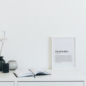 Puede incluir: Una impresi&oacute;n enmarcada en blanco con la palabra "mountains" en texto negro y una definici&oacute;n de la palabra. La impresi&oacute;n est&aacute; sobre una mesa blanca con un jarr&oacute;n negro y gris, un jarr&oacute;n de vidrio transparente y un libro cerrado.