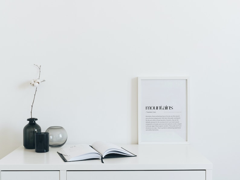 Puede incluir: Una impresi&oacute;n enmarcada en blanco con la palabra "mountains" en texto negro y una definici&oacute;n de la palabra. La impresi&oacute;n est&aacute; sobre una mesa blanca con un jarr&oacute;n negro y gris, un jarr&oacute;n de vidrio transparente y un libro cerrado.