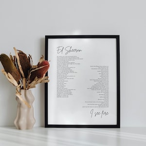 Puede incluir: Una impresión enmarcada en negro con texto blanco sobre fondo blanco. El texto dice "Ed Sheeran" e incluye la letra de la canción "I See Fire". La impresión está sobre una superficie blanca con un jarrón de flores secas delante.