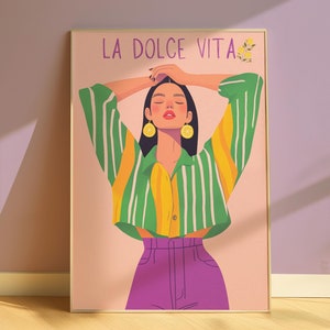 May include: A framed art print featuring a woman with her arms raised, wearing a striped green and yellow shirt, purple pants, and lemon earrings. The text "LA DOLCE VITA" is at the top. The artwork has a soft, colorful, and illustrative style.