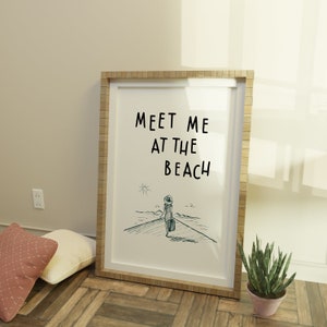 May include: A framed print with a white background and black text that reads "Meet me at the beach". The print features a simple line drawing of a person walking towards the beach with a suitcase.