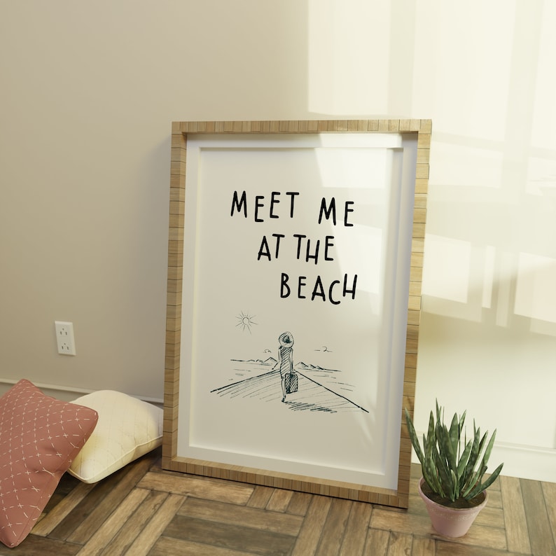 May include: A framed print with a white background and black text that reads "Meet me at the beach". The print features a simple line drawing of a person walking towards the beach with a suitcase.