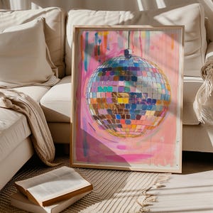 May include: A framed artwork depicting a disco ball with a multitude of mirrored squares reflecting various colours. The background is pink and abstract. The artwork is in a wooden frame and rests against a white sofa.
