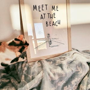 May include: A framed print with a black and white illustration of a person walking towards the beach with a suitcase. The text "Meet me at the beach" is written in black on a white background.