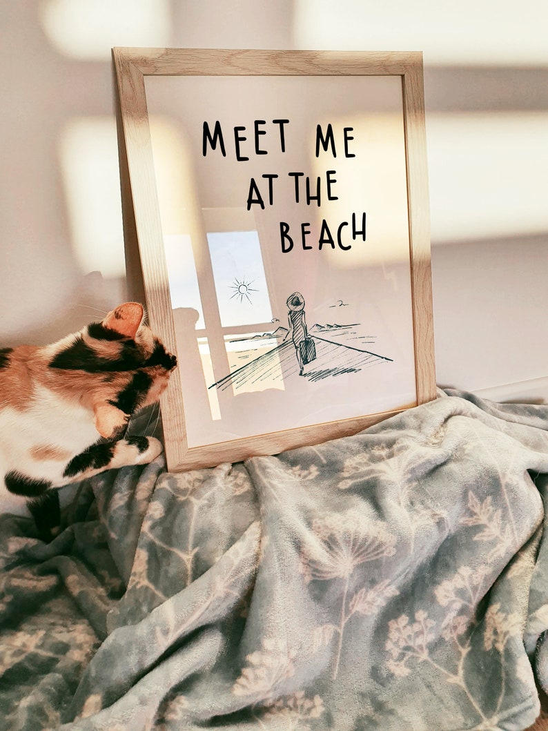May include: A framed print with a black and white illustration of a person walking towards the beach with a suitcase. The text "Meet me at the beach" is written in black on a white background.