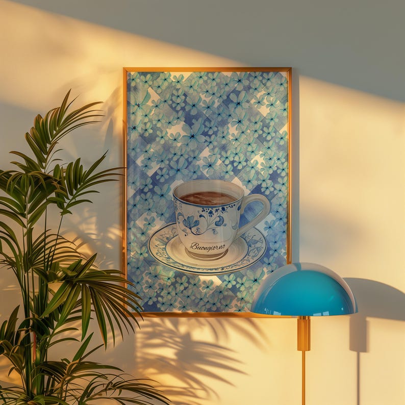 K&ouml;nnte beinhalten: Ein gerahmter Druck eines blauen und wei&szlig;en Aquarellbildes einer Tasse Kaffee mit dem Wort "Buongiorno" auf der Untertasse. Der Druck h&auml;ngt an einer wei&szlig;en Wand mit einer blauen Lampe im Vordergrund.