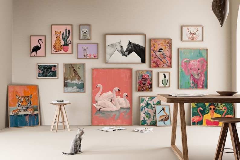 May include: A collection of framed animal illustrations in a variety of styles and colors, including a tiger, giraffe, elephant, flamingo, swan, and more. The illustrations are displayed on a white wall with a wooden table and a cat in the foreground.
