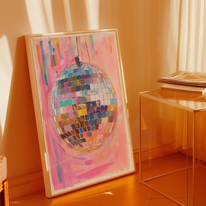 May include: A framed artwork depicting a disco ball with a multitude of coloured squares, set against a pink and blue painted backdrop. The artwork is propped against a wall in a room with warm lighting. The frame is a light colour.