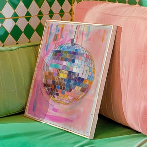 May include: A framed artwork depicting a disco ball with a multitude of colourful reflective squares, set against a pink and white painted backdrop. The frame has a gold finish. The artwork is leaning against a pink cushion.