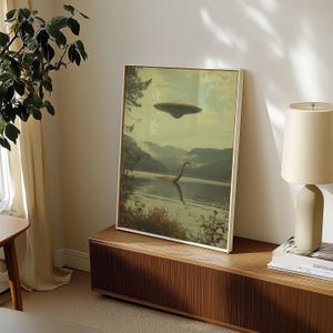 K&ouml;nnte beinhalten: Ein gerahmter Druck eines gr&uuml;nen, serpentinenartigen Wesens, das in einem See schwimmt, mit einer fliegenden Untertasse dar&uuml;ber. Der Druck ist auf einem Holzregal mit einer Lampe und einem Buch platziert.