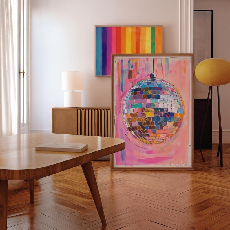 May include: A room interior with a large framed painting of a disco ball against a pink backdrop. Above, a rainbow-striped artwork is displayed. A wooden coffee table and a lamp are also present.
