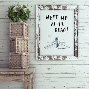 May include: Whitewashed wood wall with a white frame with the text "Meet me at the beach" and a simple line drawing of a person walking towards a lighthouse. There are three wicker baskets stacked on a wooden table.