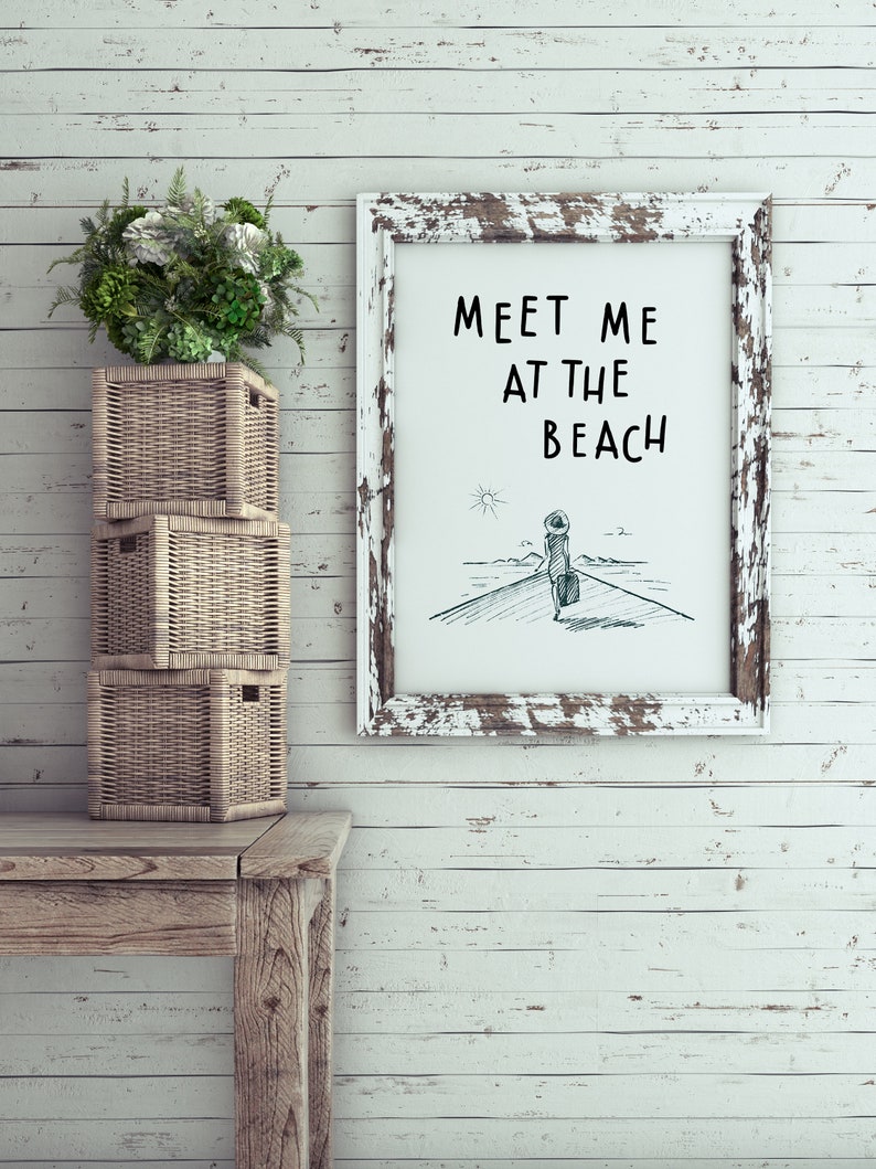 May include: Whitewashed wood wall with a white frame with the text "Meet me at the beach" and a simple line drawing of a person walking towards a lighthouse. There are three wicker baskets stacked on a wooden table.