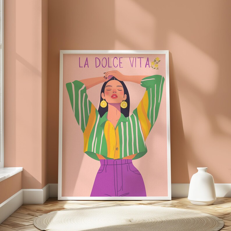 May include: A framed art print featuring a stylized illustration of a person with arms raised. The person wears a green and yellow striped shirt, purple pants, and lemon earrings. The words "LA DOLCE VITA" are at the top of the print.