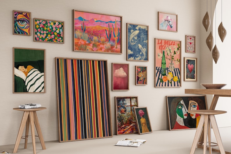 May include: A gallery wall with a variety of framed artwork, including abstract paintings, landscapes, and illustrations. The wall is painted a light beige color and the frames are all different sizes and styles. There are two small wooden stools in the foreground.
