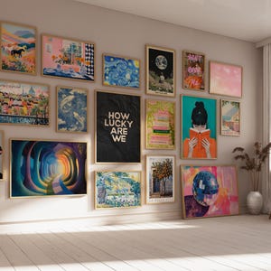 May include: A gallery wall featuring a collection of various framed art prints in light wood frames. The prints include landscapes, abstract patterns, a portrait with a pink line, a black poster with the text HOW LUCKY ARE WE, and a person reading.