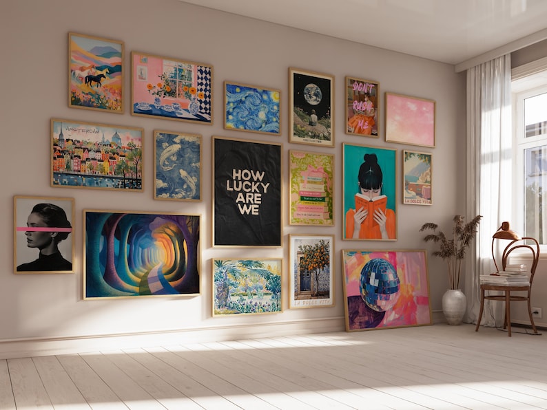 May include: A gallery wall featuring a collection of various framed art prints in light wood frames. The prints include landscapes, abstract patterns, a portrait with a pink line, a black poster with the text HOW LUCKY ARE WE, and a person reading.