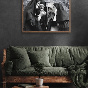 May include: Black and white photograph of four nuns standing in front of a brick wall, each holding a cigarette. The photo is framed in a brown wooden frame.