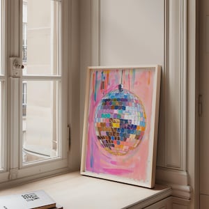 May include: A framed artwork depicting a disco ball with a vibrant, multi-coloured design set against a pink and blue backdrop. The artwork is propped on a windowsill, next to a stack of books. The disco ball is made of many squares.