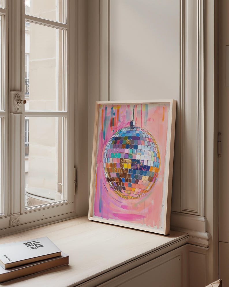 May include: A framed artwork depicting a disco ball with a vibrant, multi-coloured design set against a pink and blue backdrop. The artwork is propped on a windowsill, next to a stack of books. The disco ball is made of many squares.